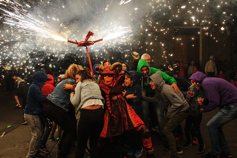 Imagen de una persona caracterizada de demonio lanzando un fuego artificial del que salen muchas chispas, al lado suyo hay personas resguardándose
