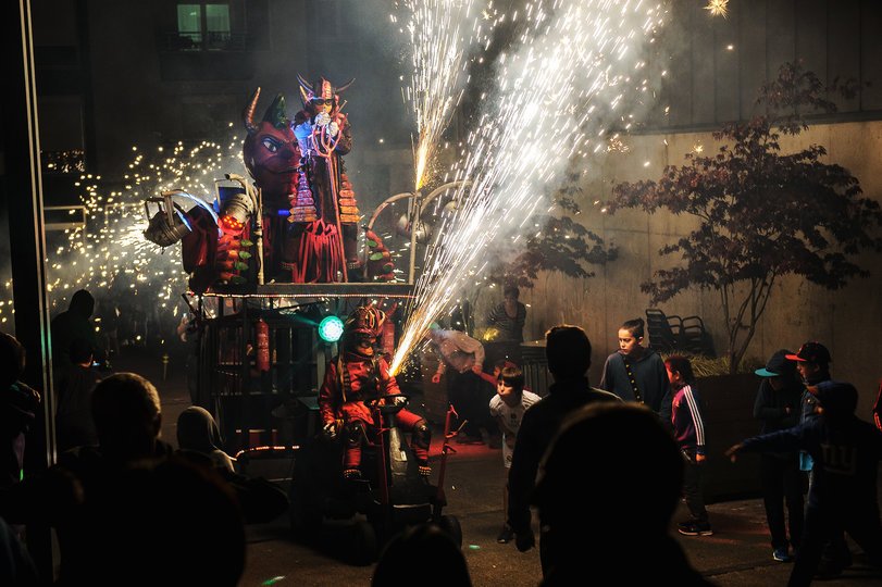 Imagen de personas caracterizadas de demonios lanzando un fuegos artificiales del que salen muchas chispas, al lado suyo hay personas
