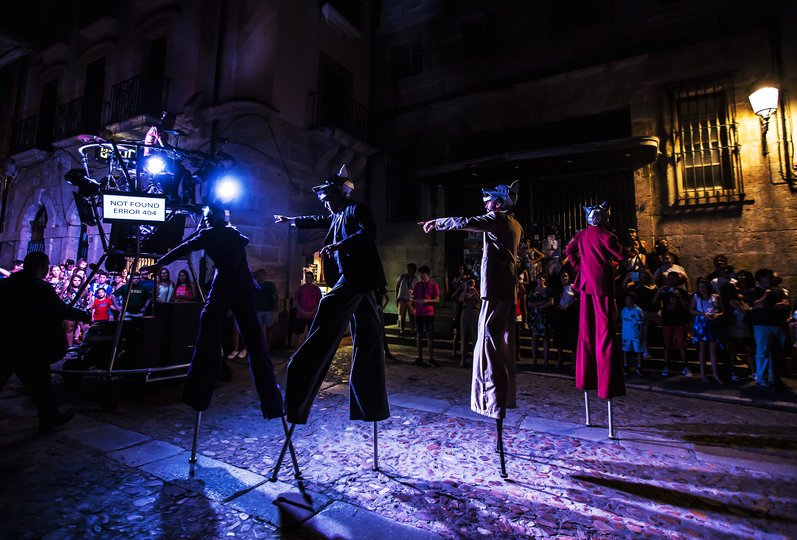 Imagen de 4 personas vestidas con trajes de colores y con máscaras de lobos en frente de público de noche e iluminados por varios focos