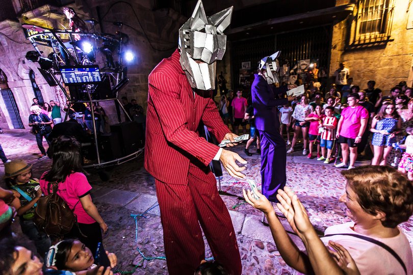 Imagen de una persona vertida con un traje rojo con máscara de lobo repartiendo dinero a una señora